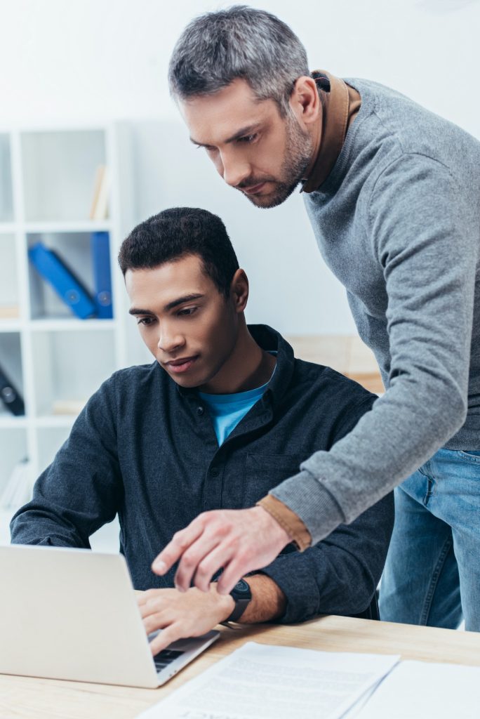 bearded business mentor looking at young colleague using laptop in office